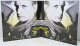A folded book or album cover featuring a black-and-white portrait of a man wearing a striped shirt, with a city skyline at the bottom and the title 'How to Become the Cat' in yellow text.
