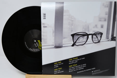 A vinyl record partially pulled from its sleeve, showing the black record and the back cover artwork with a black-and-white photo of eyeglasses by a window and track listing text.