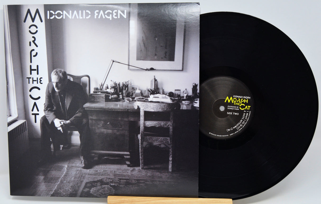 Donald Fagen's 'Morph the Cat' vinyl record LP, showing the black-and-white album cover sleeve on the left and the black vinyl record partially pulled out on the right, resting on a light wood stand against a white background.