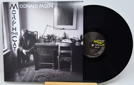 Donald Fagen's 'Morph the Cat' vinyl record LP, showing the black-and-white album cover sleeve on the left and the black vinyl record partially pulled out on the right, resting on a light wood stand against a white background.