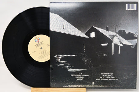 A vinyl record partially pulled from its sleeve, showing the black record on the left and the album cover on the right. The cover features a black-and-white photograph of a house and lists track titles including 'Green Flower Street' and 'New Frontier.
