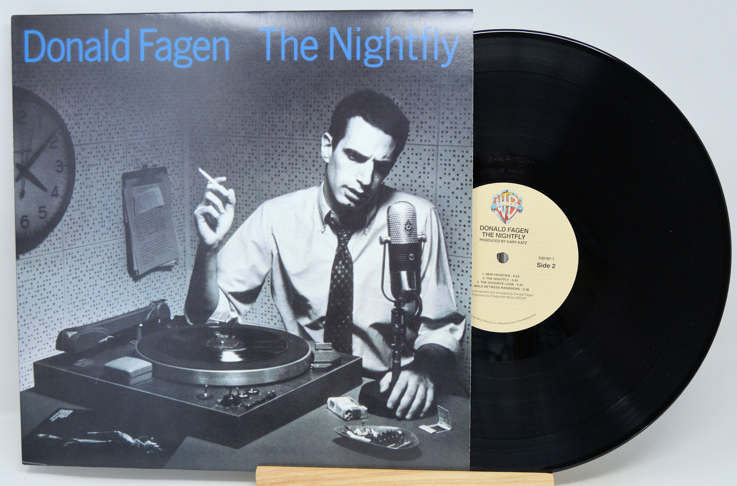 Donald Fagen's 'The Nightly' vinyl record album with the LP partially pulled from the jacket, displayed on a small wooden stand against a light gray background.