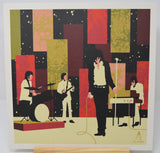 A square art print featuring a stylized band performance scene with four musicians against an abstract geometric background in black, red, olive green, and beige. The print is shown standing on a light wood surface with a white border around the artwork.