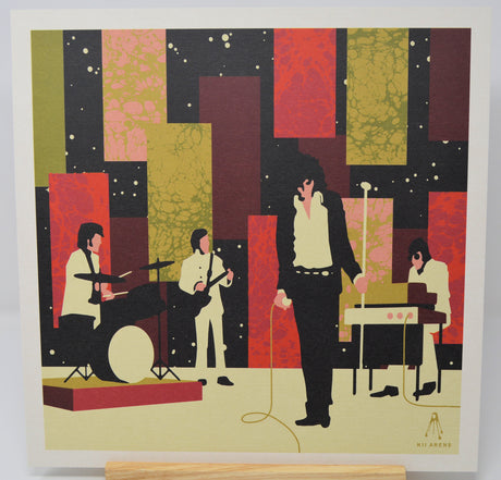 A square art print featuring a stylized band performance scene with four musicians against an abstract geometric background in black, red, olive green, and beige. The print is shown standing on a light wood surface with a white border around the artwork.