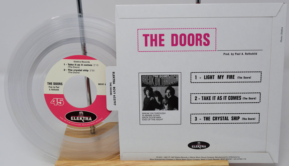 A boxed The Doors 45 rpm vinyl record set on a wooden display stand, showing the white outer sleeve with pink 'THE DOORS' text and track list, alongside the clear vinyl record with a pink and white center label.