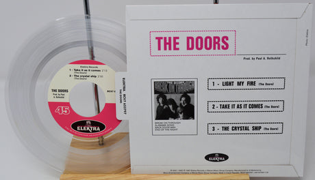 A boxed The Doors 45 rpm vinyl record set on a wooden display stand, showing the white outer sleeve with pink 'THE DOORS' text and track list, alongside the clear vinyl record with a pink and white center label.