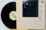 A vinyl record partially pulled from its back cover sleeve, photographed on a light background. The black record has a cream label, and the sleeve features a dark photo of a person playing an instrument and printed track listing and text blocks.