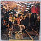 The Basement Tapes 'Original Master Recording' vinyl LP cover, sealed in its plastic shrink wrap and displayed on a wooden stand. The cover art features a crowded, vintage recording studio scene with musicians playing accordions, guitars, and a harmonica, with a small dog in the foreground.