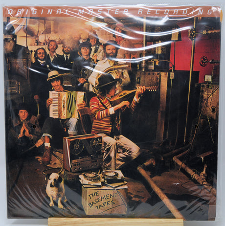 The Basement Tapes 'Original Master Recording' vinyl LP cover, sealed in its plastic shrink wrap and displayed on a wooden stand. The cover art features a crowded, vintage recording studio scene with musicians playing accordions, guitars, and a harmonica, with a small dog in the foreground.