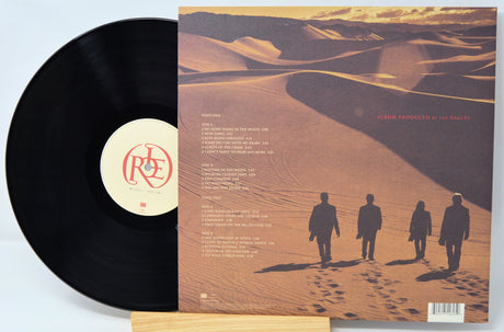 A vinyl record partially pulled from its album cover, displayed on a small wooden stand against a light gray background. The record is black with a cream center label featuring red lettering.