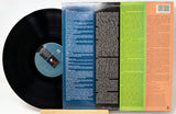 A vinyl record partially pulled from its back cover, showing a black record on the left and a colorful printed back cover with blue, black, green, and peach sections on the right.