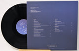 A vinyl record partially pulled from its sleeve, showing the black record with a blue label next to the dark blue printed inner sleeve with track listings.