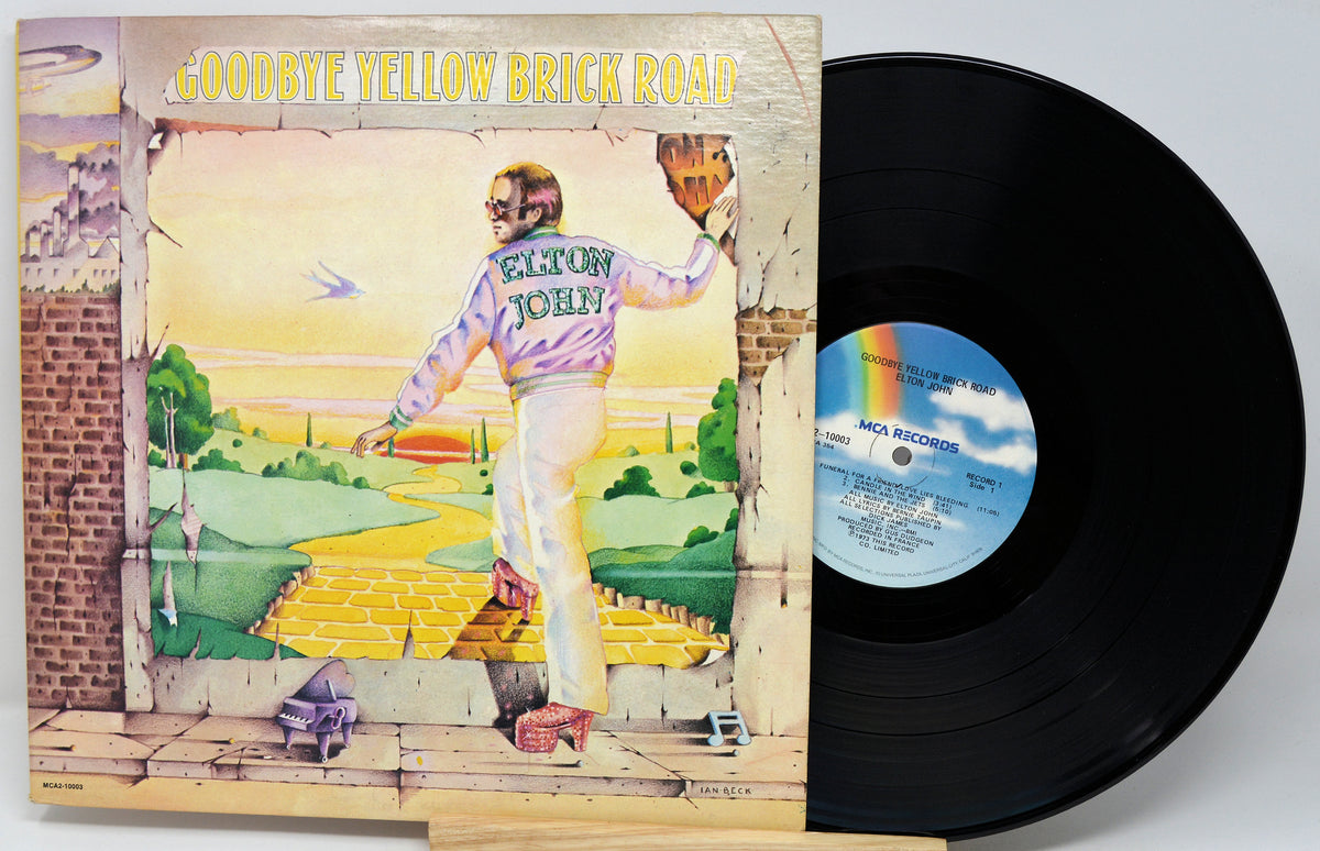 Elton John's 'Goodbye Yellow Brick Road' vinyl record LP with its illustrated album cover partially covering the black record, photographed on a light neutral background.
