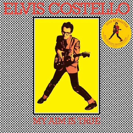 Elvis Costello 'My Aim Is True' album cover artwork featuring a stylized portrait of the artist playing an electric guitar against a bright yellow background, framed by a black-and-white checkered border with large red text reading 'ELVIS COSTELLO' at the top and 'MY AIM IS TRUE' at the bottom. A yellow circular 'Road Warrior' sticker graphic appears on the right.