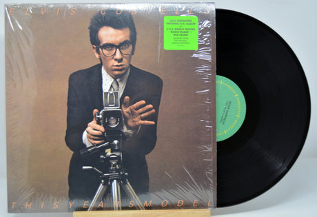 Vinyl record with a cover featuring Elvis Costello holding a camera, on a brown background.