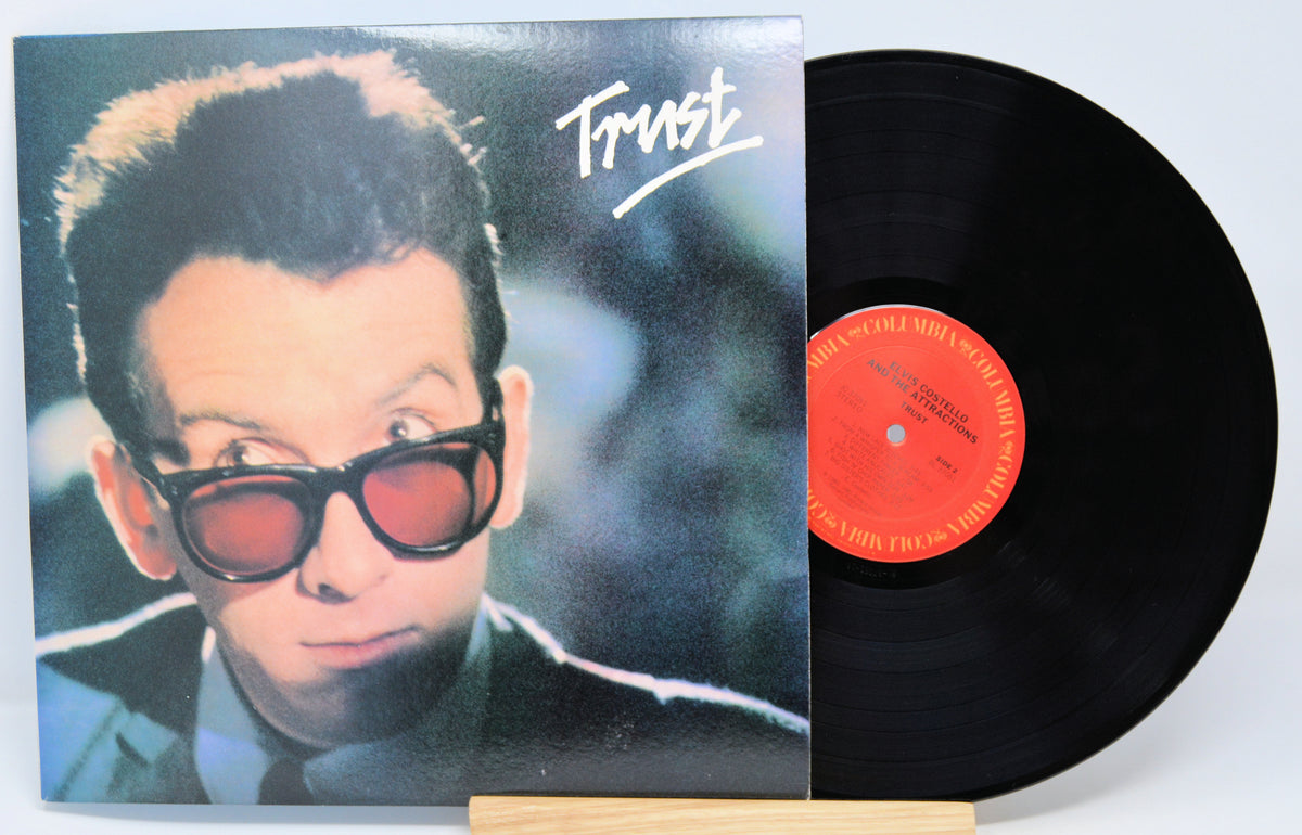 Elvis Costello vinyl record album 'Trust' with a picture of Elvis Costello wearing sunglasses on the cover, beside the vinyl disc.