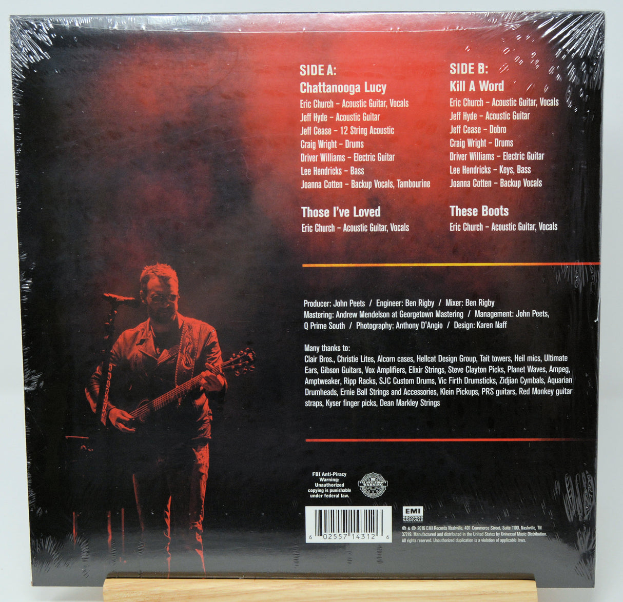 The back cover of a vinyl record album wrapped in clear plastic, displayed on a small wooden stand. The cover features a dark red and black background with a faint live performance photo, listing track titles and artist credits for Chattanooga Lucy, Kill A Word, These I've Loved, and These Boots.