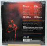 The back cover of a vinyl record album wrapped in clear plastic, displayed on a small wooden stand. The cover features a dark red and black background with a faint live performance photo, listing track titles and artist credits for Chattanooga Lucy, Kill A Word, These I've Loved, and These Boots.