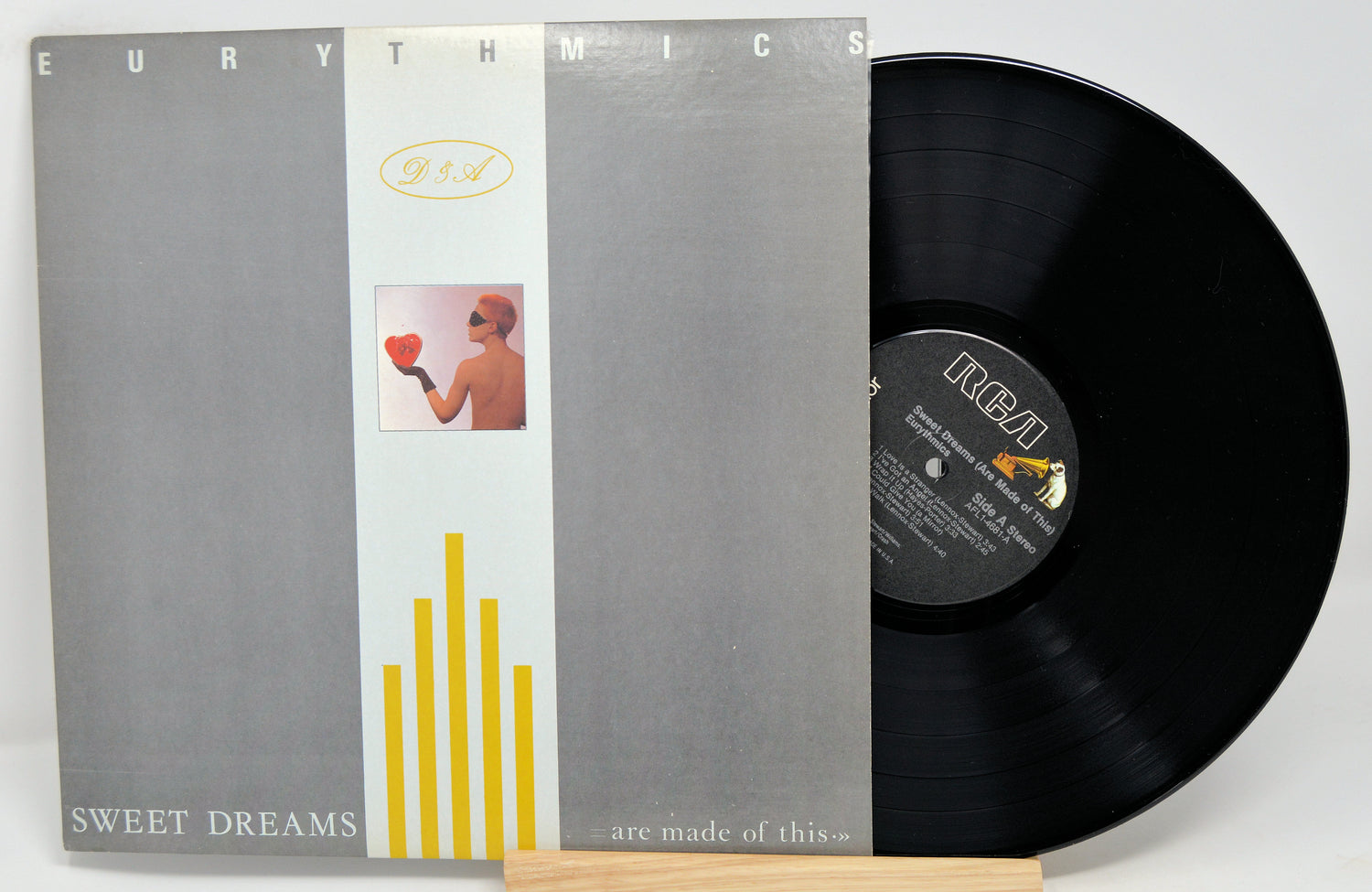 Eurythmics - Sweet Dreams album cover and vinyl record. The cover features a gray background with yellow and white stripes and a silhouette of a person holding a heart.