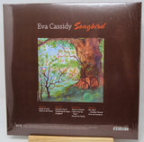 Eva Cassidy 'Songbird' vinyl record album cover, shown in shrink wrap on a wooden display stand. The cover is dark brown with the artist's name and album title in white and orange text, featuring a colorful painting of a tree trunk and branches.