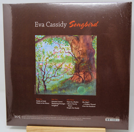 Eva Cassidy 'Songbird' vinyl record album cover, shown in shrink wrap on a wooden display stand. The cover is dark brown with the artist's name and album title in white and orange text, featuring a colorful painting of a tree trunk and branches.