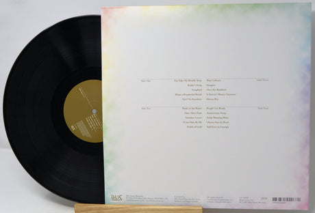 A vinyl record partially pulled from its back cover, showing the tracklist and pastel rainbow watercolor-style border on the sleeve, photographed on a light background.