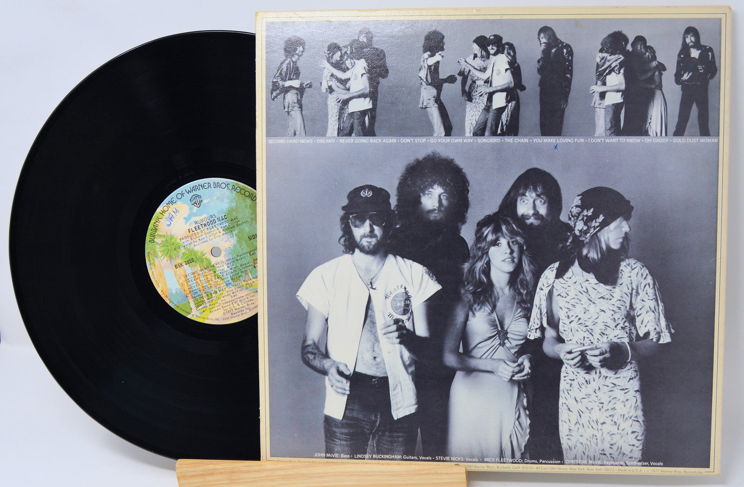 A black vinyl record partially pulled from its sleeve, showing the front cover artwork with a black-and-white band photo and a small multicolor label on the record, displayed on a wooden stand against a white background.