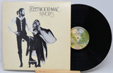 Fleetwood Mac 'Rumours' vinyl record album with the illustrated sleeve cover partially covering a black LP record on a light gray background.