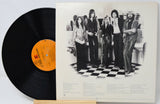 A vinyl record partially pulled from its printed sleeve, showing a black record with a bright orange center label and the back cover featuring a black-and-white band photo on a cream background.