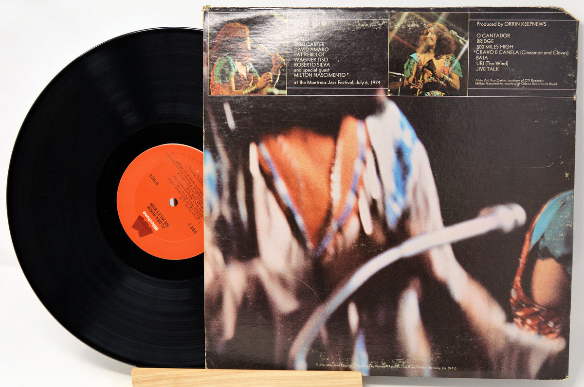 A vinyl record partially pulled from its album sleeve, photographed on a light background. The black record has an orange center label, and the sleeve features a dark cover with a close-up photo and printed track listings.