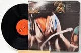 A vinyl record partially pulled from its album sleeve, photographed on a light background. The black record has an orange center label, and the sleeve features a dark cover with a close-up photo and printed track listings.