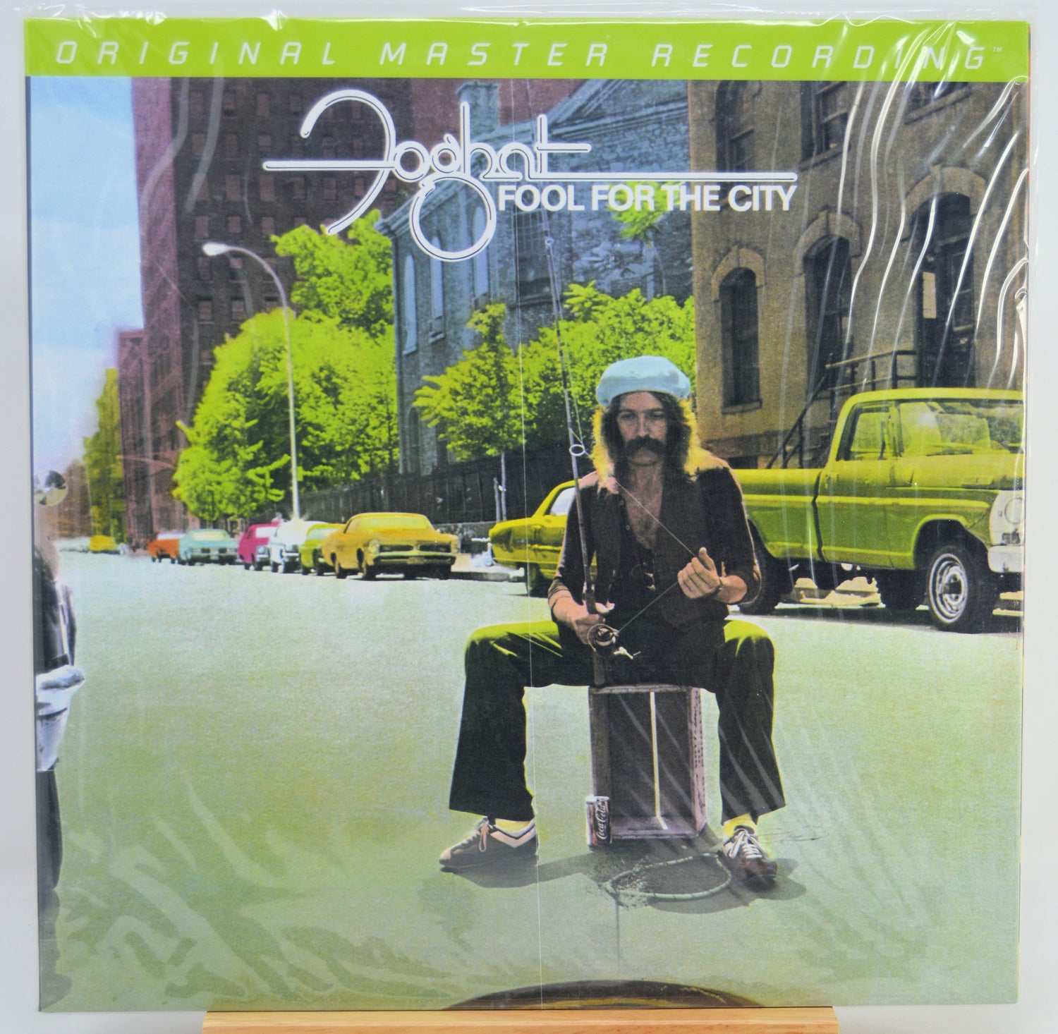 The album cover of Foghat's 'Fool for the City' on a vinyl record, sealed in its plastic shrink wrap. The cover art features a surreal street scene with a bearded man sitting on a drum in the middle of the road, flanked by yellow taxis and green cars, with the artist's name and album title prominently displayed at the top.