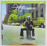 The album cover of Foghat's 'Fool for the City' on a vinyl record, sealed in its plastic shrink wrap. The cover art features a surreal street scene with a bearded man sitting on a drum in the middle of the road, flanked by yellow taxis and green cars, with the artist's name and album title prominently displayed at the top.