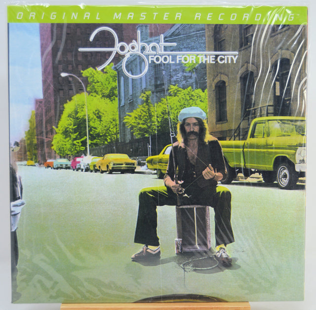 The album cover of Foghat's 'Fool for the City' on a vinyl record, sealed in its plastic shrink wrap. The cover art features a surreal street scene with a bearded man sitting on a drum in the middle of the road, flanked by yellow taxis and green cars, with the artist's name and album title prominently displayed at the top.