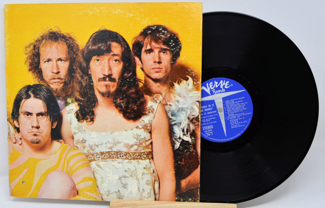 A vinyl record partially pulled from its sleeve, showing the band members on the cover art against a yellow background, with the black record and blue label visible on a light gray background.