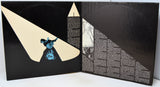 An open book or album cover spread showing a black background with cream geometric shapes, a small blue figure illustration, and dense white text blocks.