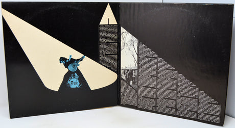 An open book or album cover spread showing a black background with cream geometric shapes, a small blue figure illustration, and dense white text blocks.