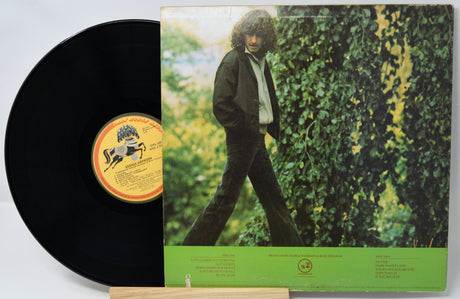Vinyl record with a cover featuring a person standing in front of a green hedge.