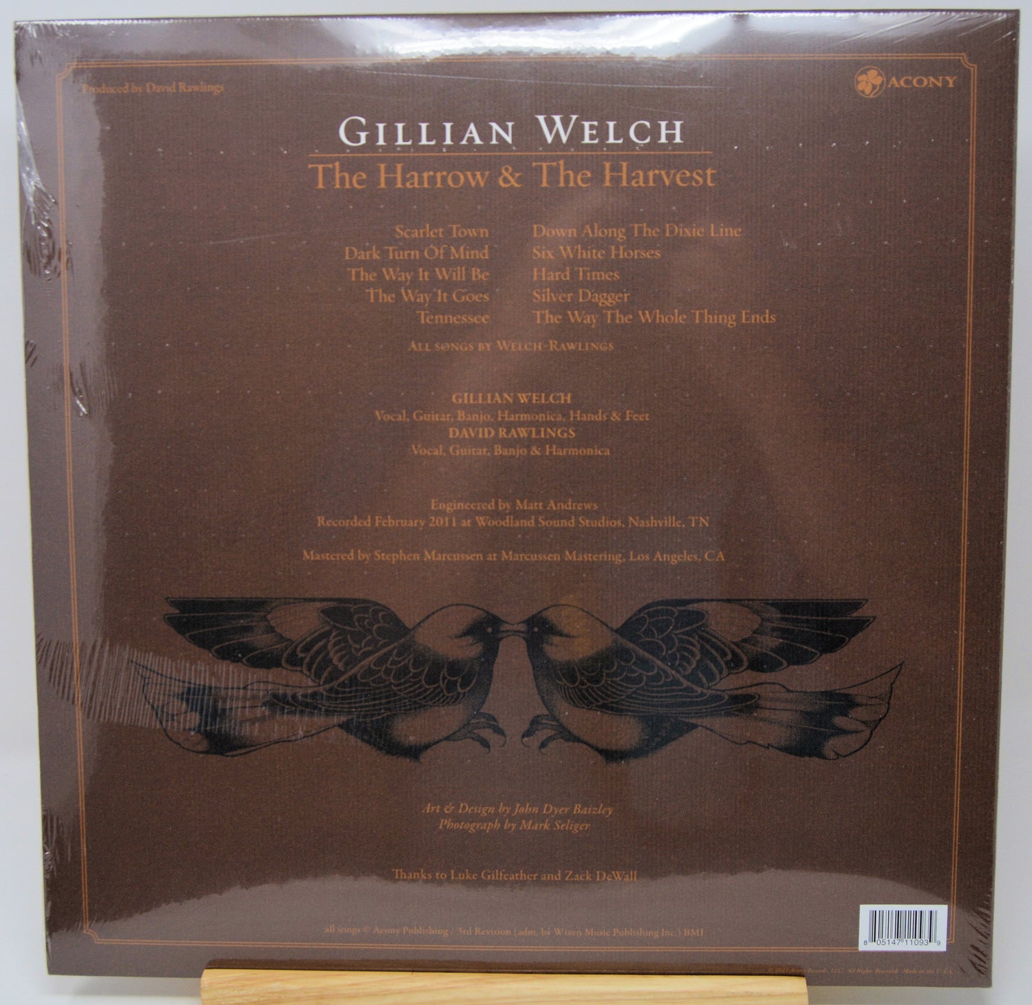 The back cover of Gillian Welch's 'The Harrow & The Harvest' vinyl record album on Acoustic Records, featuring a dark brown background with tracklist text and a black bird illustration.
