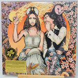 The album cover of 'The God Machine' by the band 'The Holy Ghost', displayed on a wooden stand. The artwork features a stylized illustration of a woman in a light green corset and a man in a white shirt and blue jeans, surrounded by a fiery orange ring, a owl, and autumn foliage.