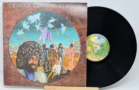 Vinyl record with colorful cover featuring a group of people and a central figure, on a light gray background.