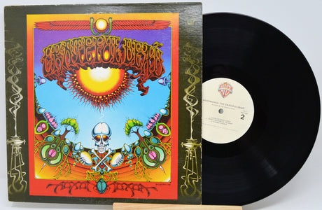 A vinyl record album shown with its illustrated outer sleeve and the black record partially pulled out. The cover art features a psychedelic sun, skull, and ornate decorative motifs in bright orange, yellow, blue, green, and red tones.