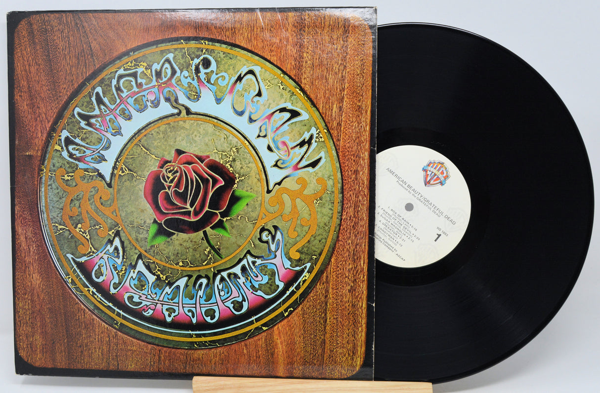 A vinyl record album with its illustrated cover jacket standing beside the black record on a small wooden display stand against a light gray background.
