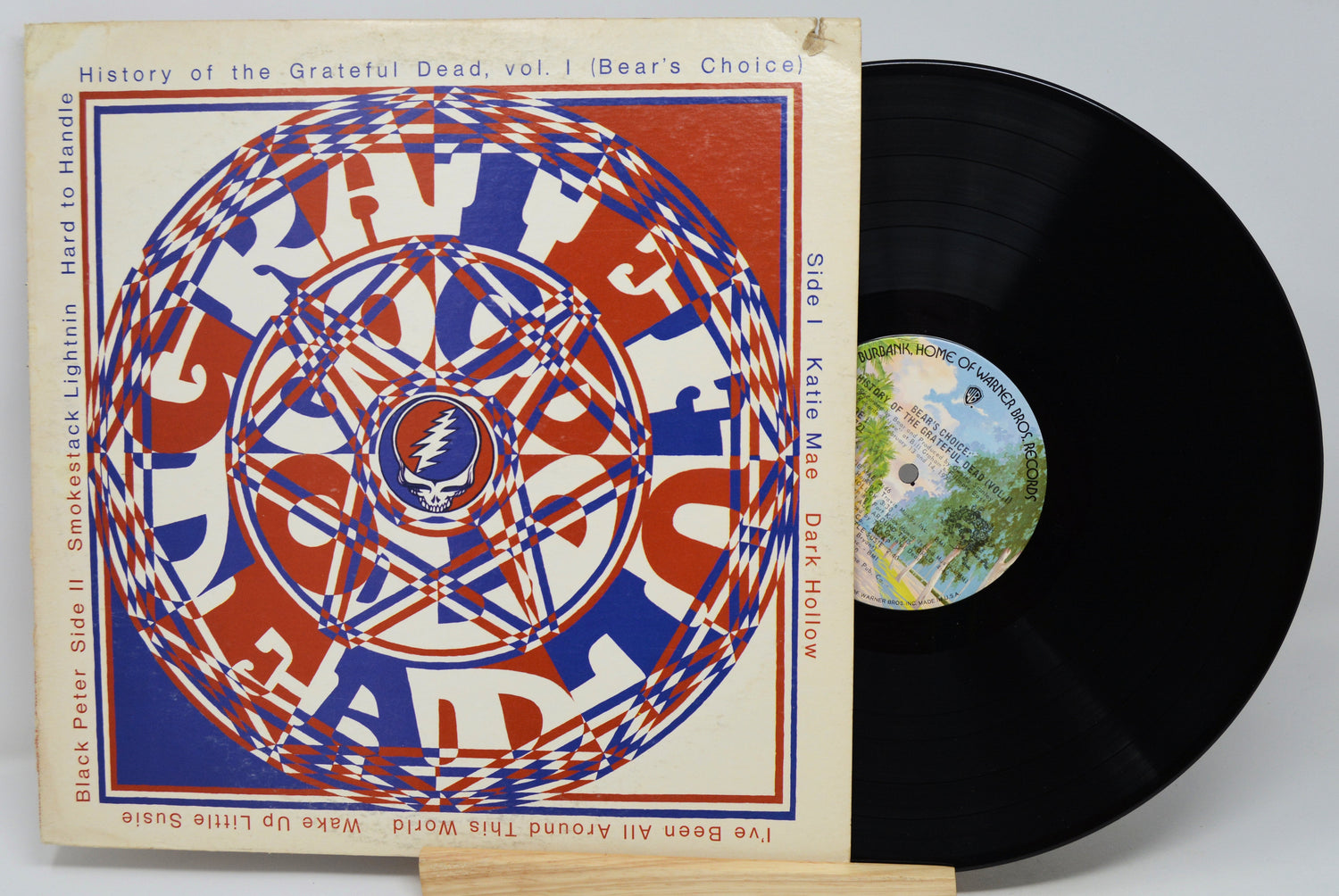 Grateful Dead 'History of the Grateful Dead, vol. 1' vinyl record LP with its illustrated sleeve and the black record partially visible, displayed on a small stand against a light gray background.