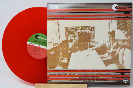 A vinyl record partially pulled from its album cover, showing a bright red translucent record and a printed cover with a sepia-toned photo and red-and-white striping. The record label reads Polydor.