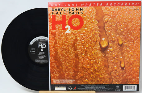 Vinyl record album shown with the black record partially pulled from the cover, which features the title 'H2O' and artist Daryl John Hall Oates on a textured orange background with water droplets.