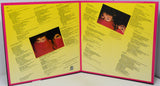 An open vinyl record sleeve or booklet with bright yellow printed pages, pink borders, and two small inset photos of a person in red clothing, photographed against a plain light background.