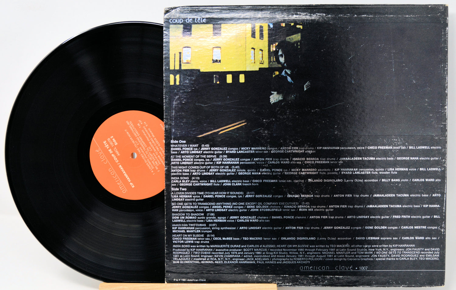 A vinyl record partially pulled from its worn black outer sleeve, showing the orange center label and the back cover artwork with a yellow building and dense white text on a black background.