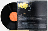 A vinyl record partially pulled from its worn black outer sleeve, showing the orange center label and the back cover artwork with a yellow building and dense white text on a black background.
