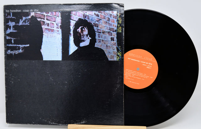 A vinyl record partially pulled from its sleeve, showing the black LP disc and the album cover. The cover features a black-and-white photo of a person in front of a brick wall, with a bright orange center label.